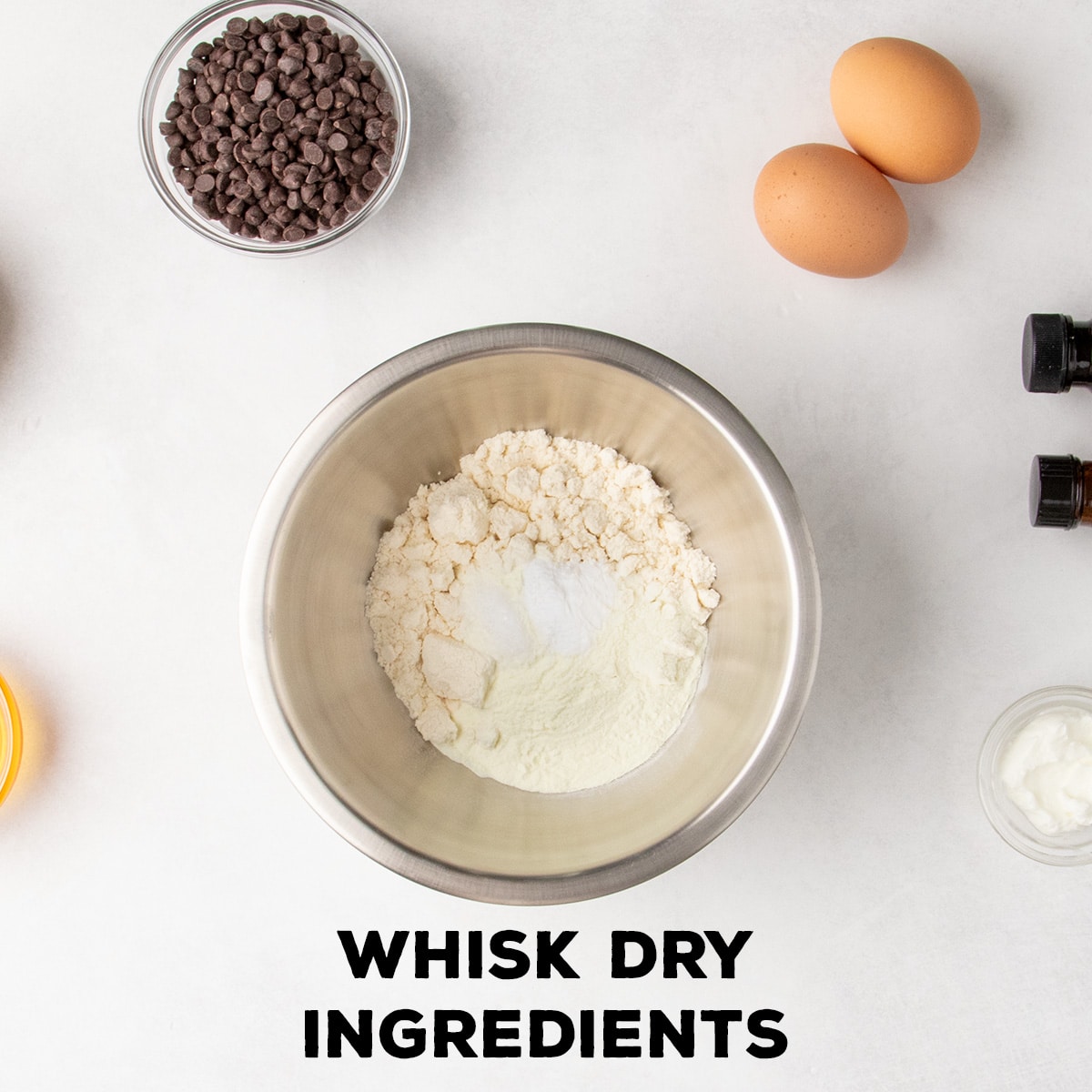 A metal mixing bowl holds several pale dry ingredients, with eggs, mini chocolate chips, yogurt, and bottles arranged around it; text reads “WHISK DRY INGREDIENTS.”