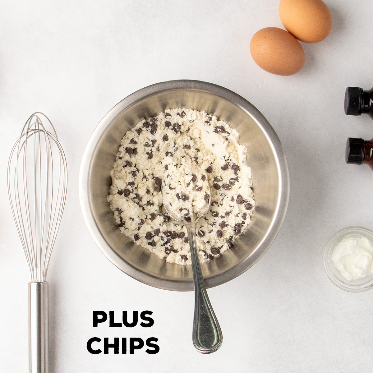 Mini chocolate chips are mixed into pale dry ingredients in a metal bowl with a spoon; text reads “PLUS CHIPS.”