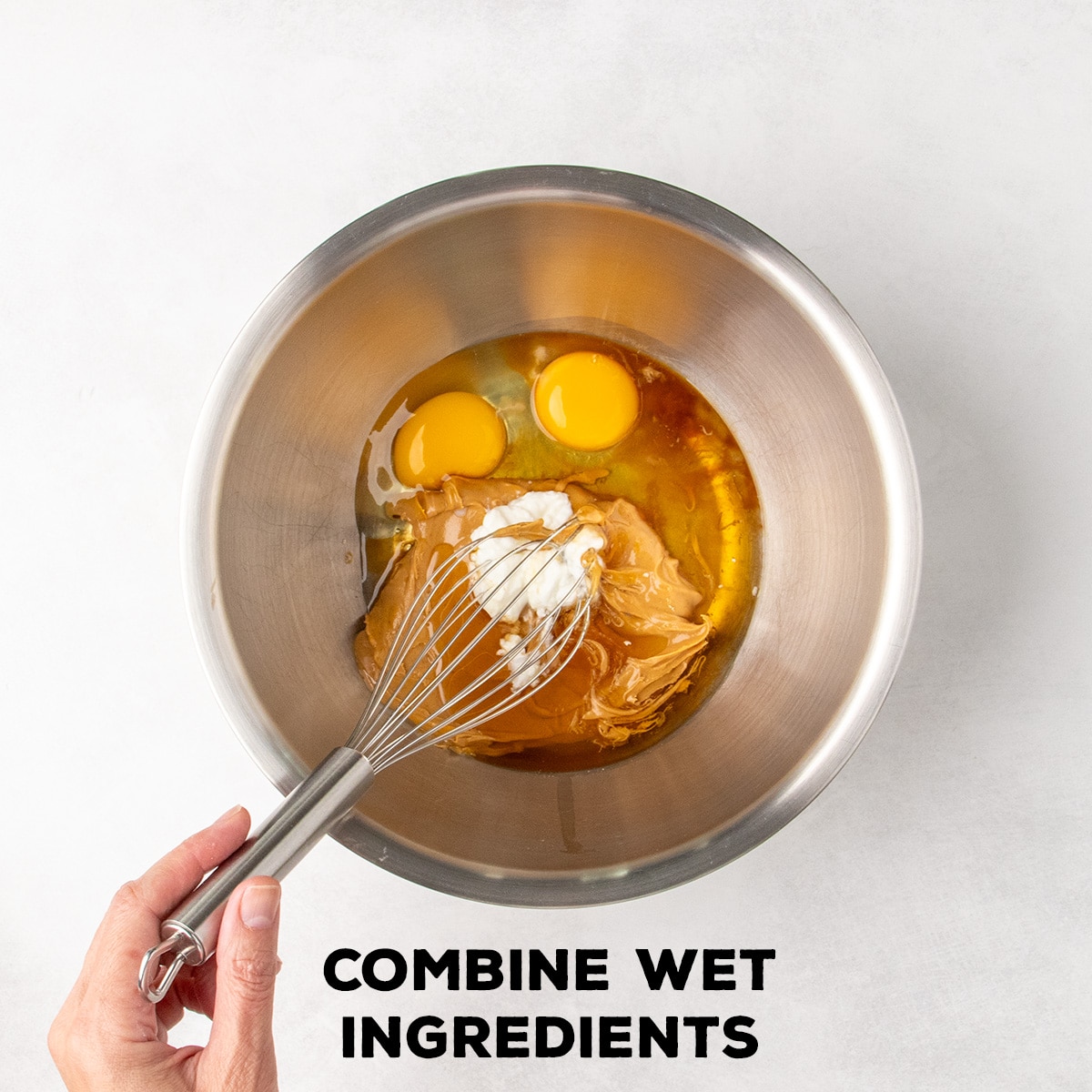 Eggs, peanut butter, yogurt, and amber liquid sit together in a metal mixing bowl as a hand holds a whisk; text reads “COMBINE WET INGREDIENTS.”