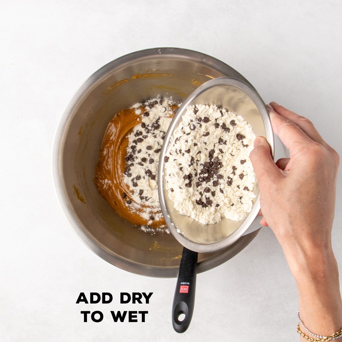 A hand tips a bowl of dry ingredients and mini chocolate chips into the wet mixture below; text reads “ADD DRY TO WET.”