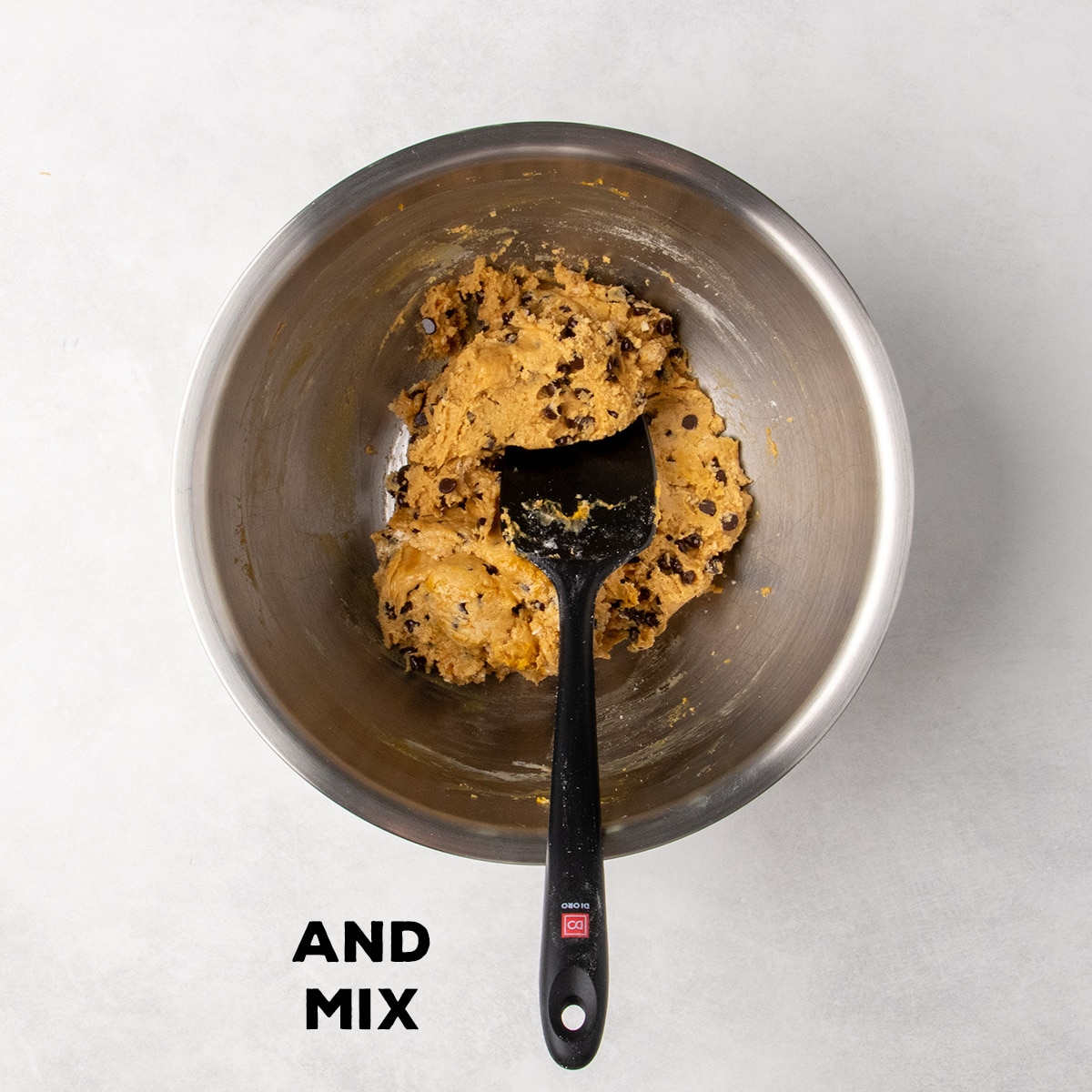 Thick cookie dough studded with mini chocolate chips sits in a metal mixing bowl with a black spatula; text reads “AND MIX.”