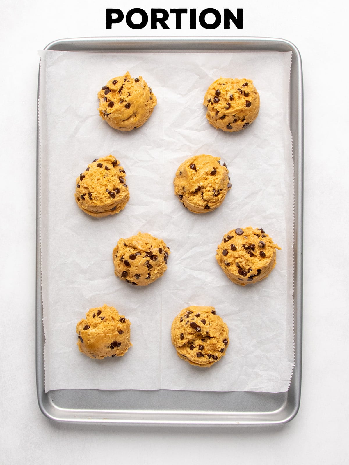 Eight scoops of light brown raw cookie dough sit spaced apart on a parchment-lined baking sheet; text reads “PORTION.”