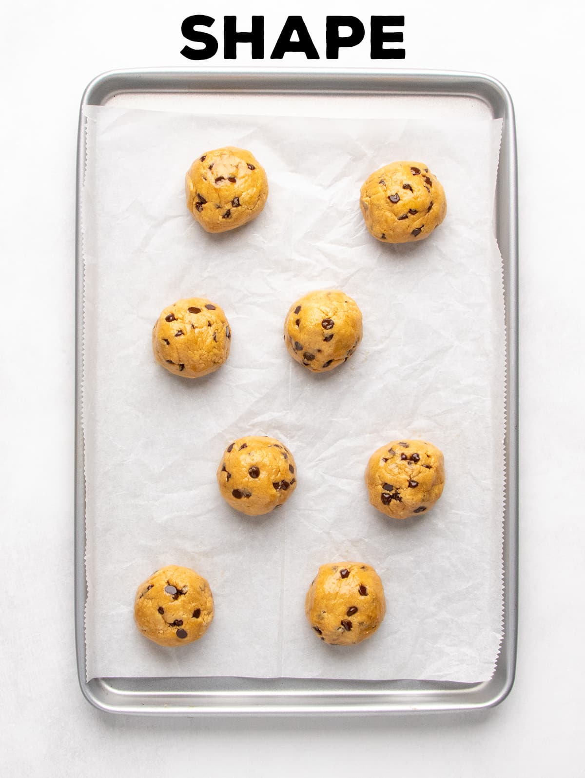 Eight rounded balls of light brown raw cookie dough with mini chocolate chips sit on a parchment-lined baking sheet; text reads “SHAPE.”