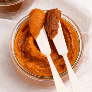 Closeup image of two white spreaders holding scoops of orange and dark orange-brown pumpkin butter over a glass jar filled with orange pumpkin butter.