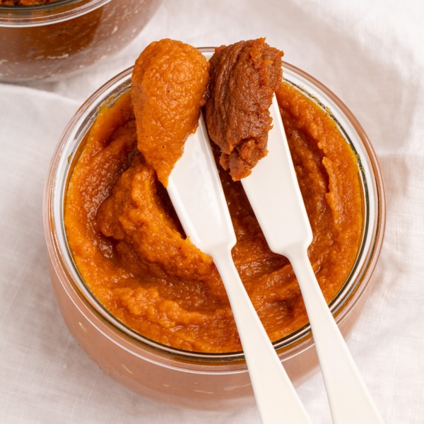 Closeup image of two white spreaders holding scoops of orange and dark orange-brown pumpkin butter over a glass jar filled with orange pumpkin butter.
