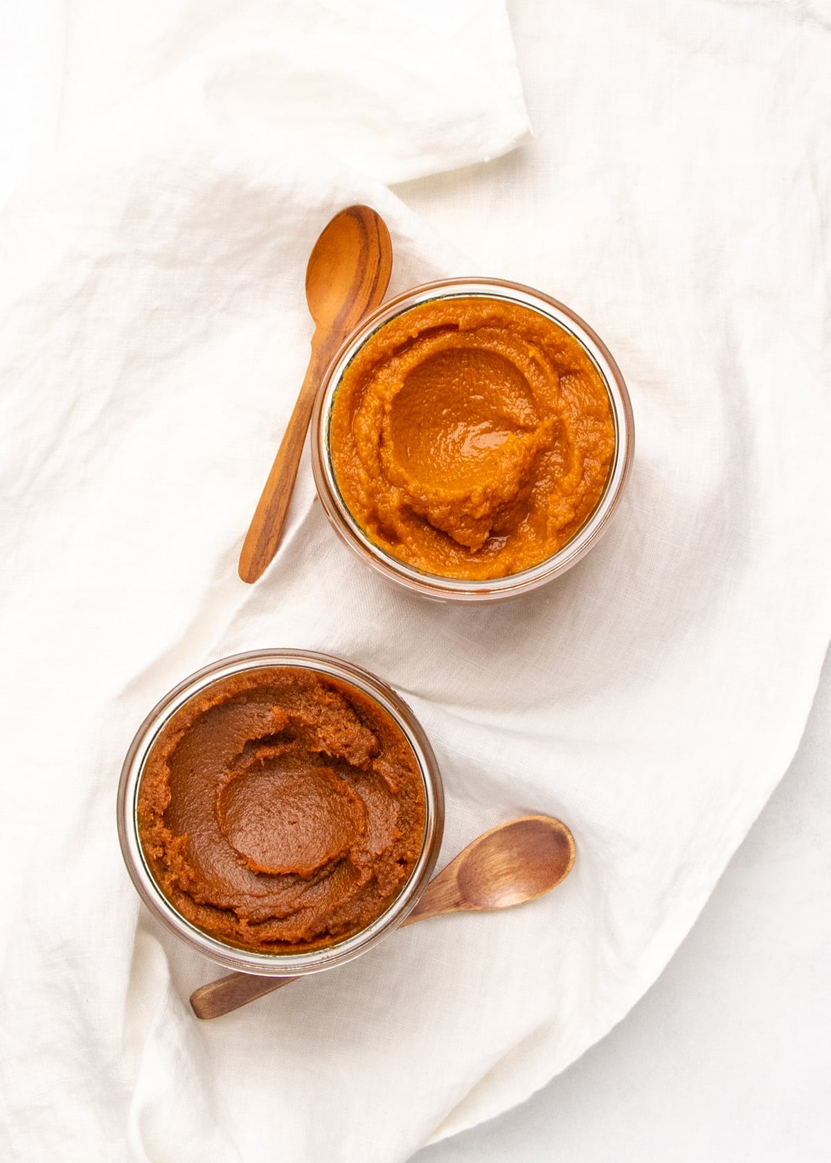Two glass jars hold orange and dark orange-brown pumpkin butter on a white cloth, with wooden spoons beside them.