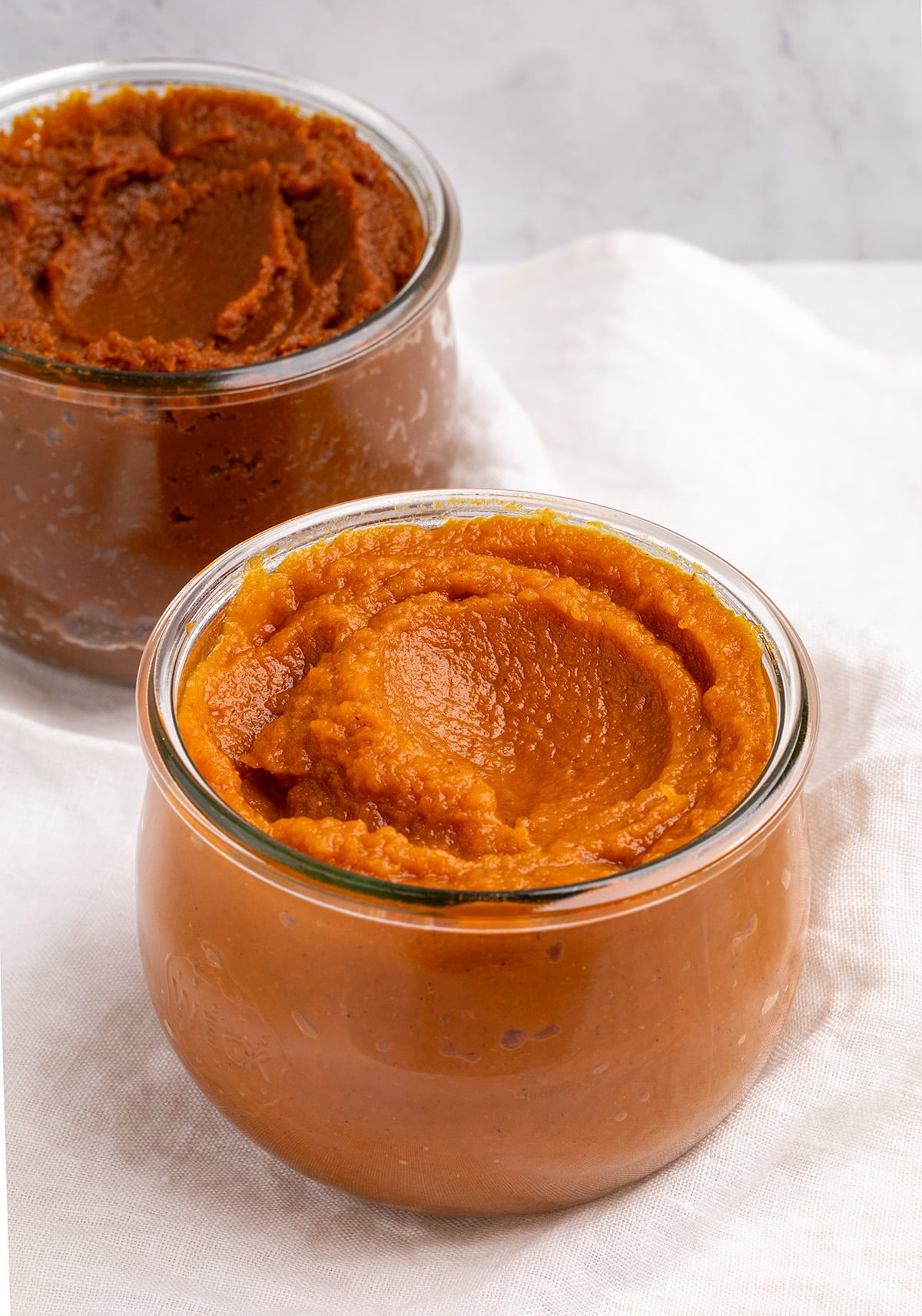 A glass jar filled with orange pumpkin butter sits in front of a second jar filled with dark orange-brown pumpkin butter on a white cloth.