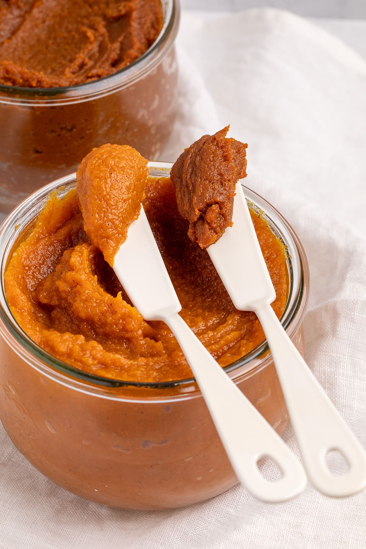 Two white spreaders hold scoops of orange and dark orange-brown pumpkin butter over a glass jar of orange pumpkin butter, with a darker jar behind it.