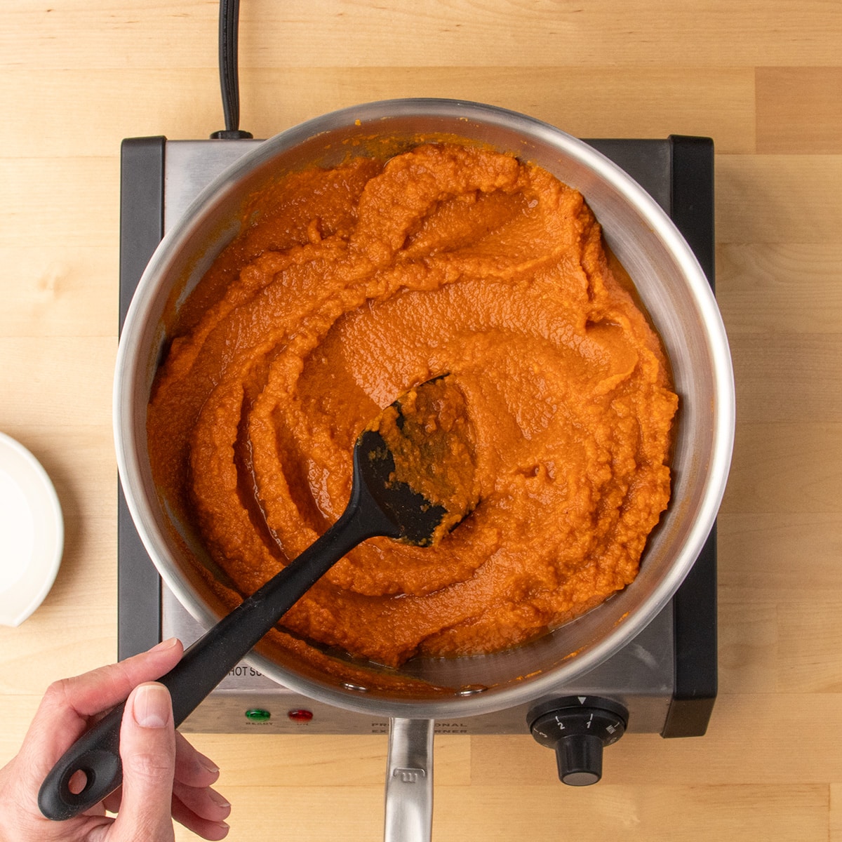 A hand stirs a thick orange pumpkin mixture in a stainless steel pan with a black silicone spatula.