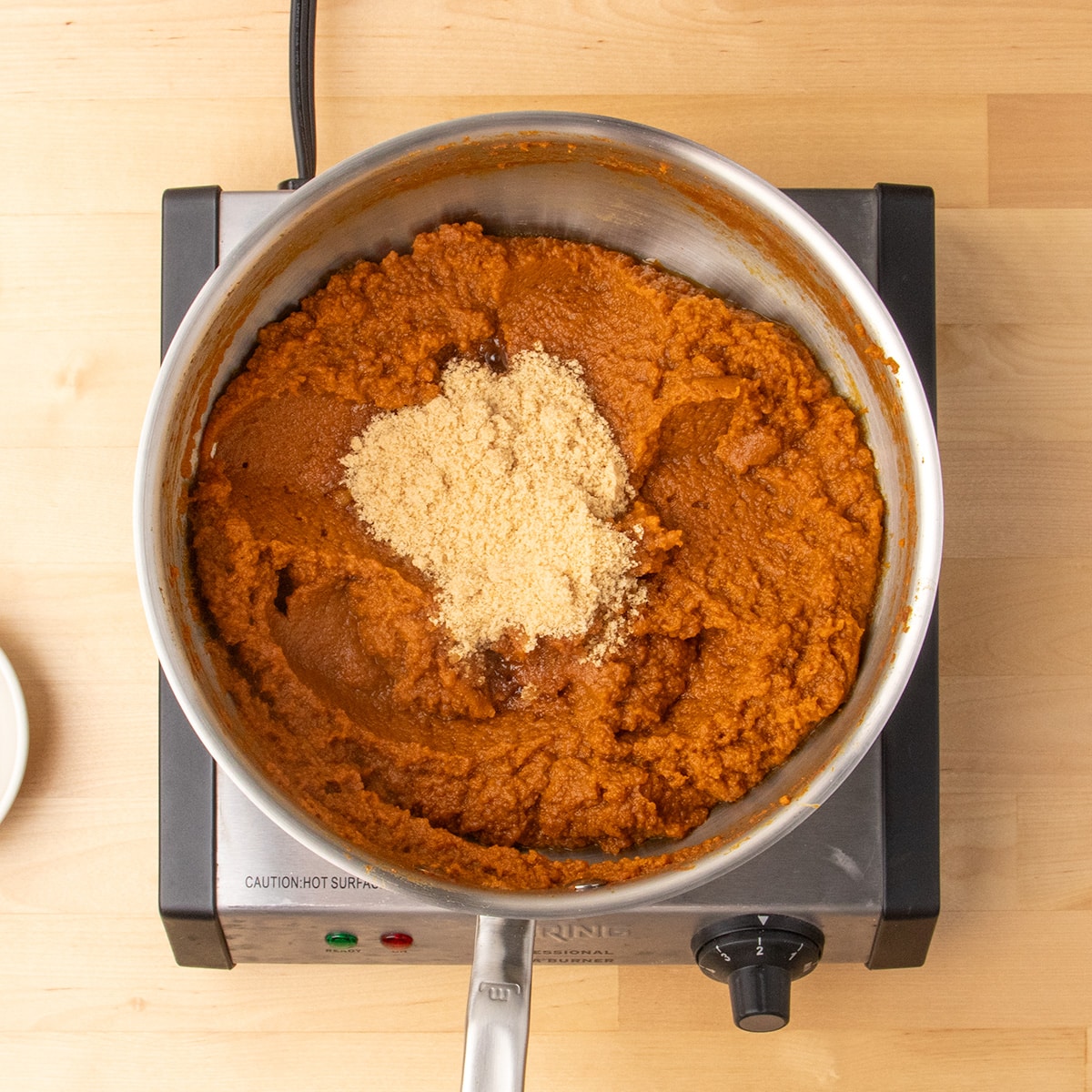 A mound of light brown granulated ingredient sits on top of a thick orange pumpkin mixture in a stainless steel pan.