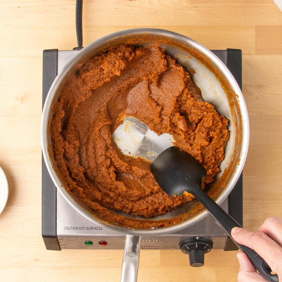 A hand stirs a dark orange, thickened pumpkin mixture in a stainless steel pan with a black spatula that's exposing the bare bottom of the pan in the center.