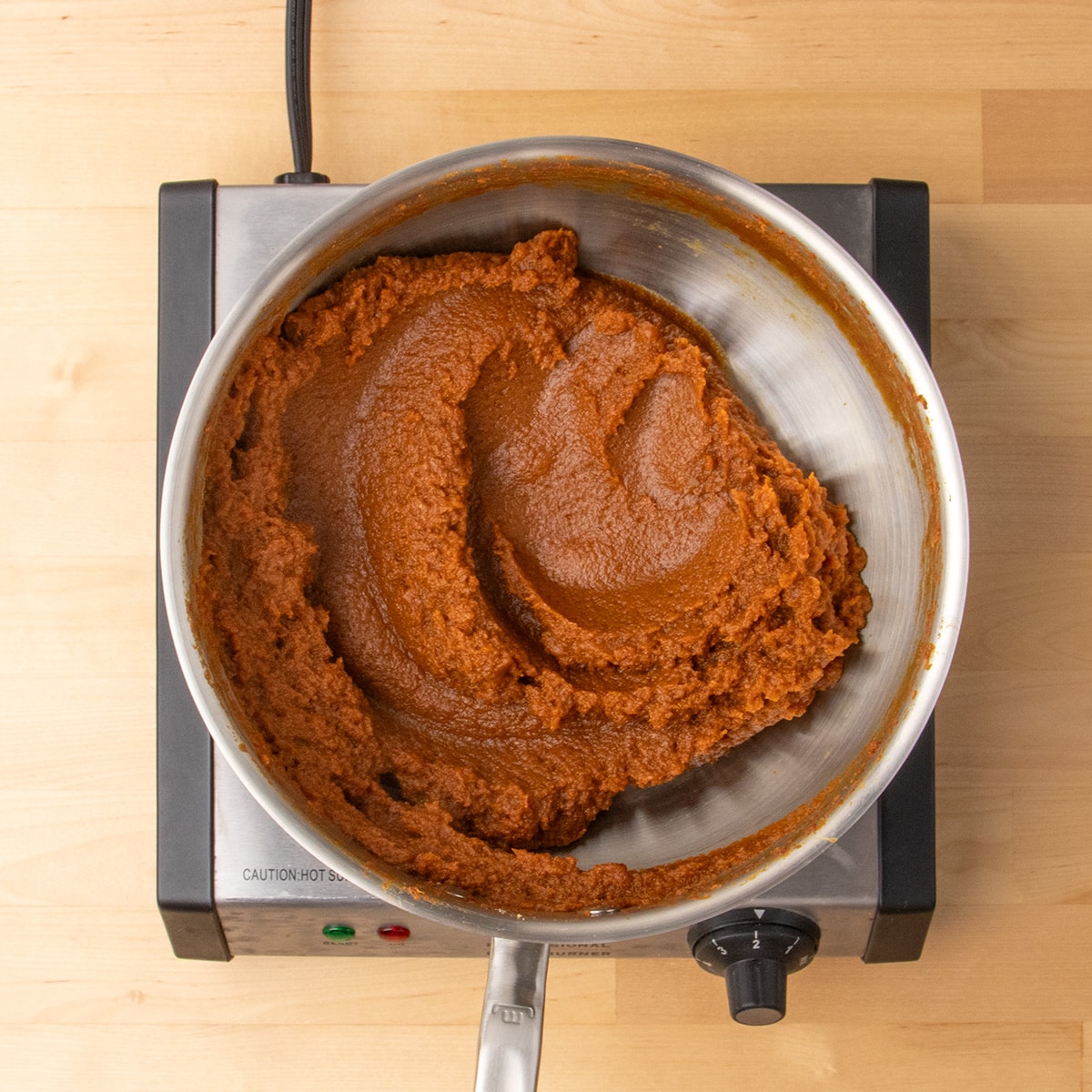 A thick, dark orange pumpkin mixture sits in a stainless steel pan on an electric burner.