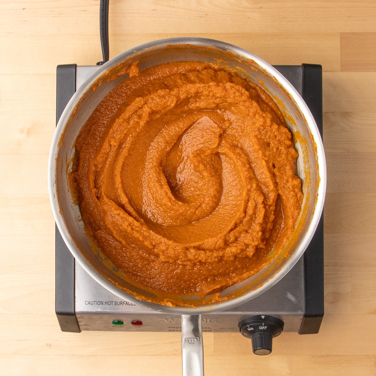 A thick but glossy lighter orange pumpkin mixture fills a stainless steel pan on an electric burner.