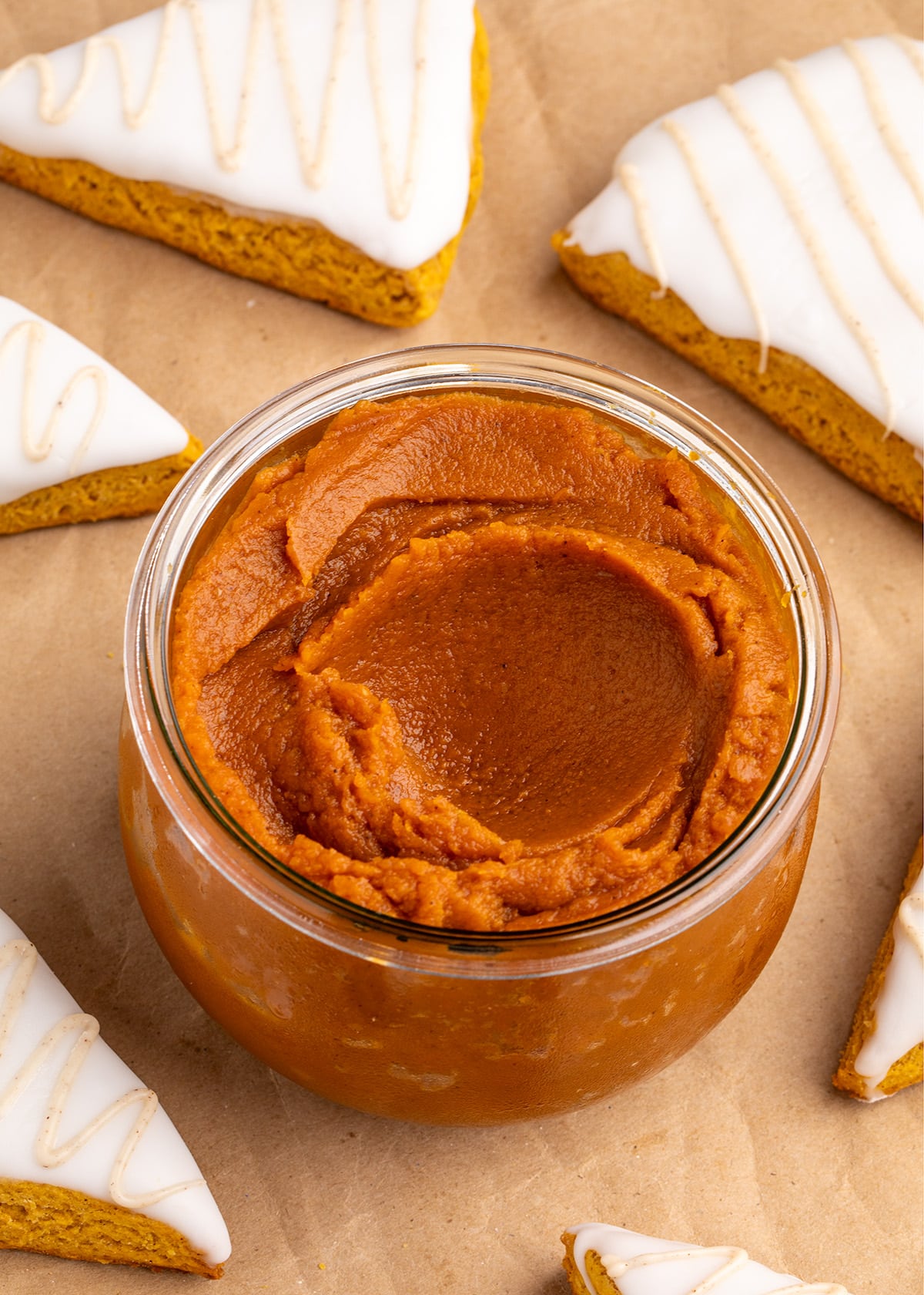 A glass jar filled with pumpkin butter sits surrounded by iced triangular pumpkin scones on brown parchment paper.