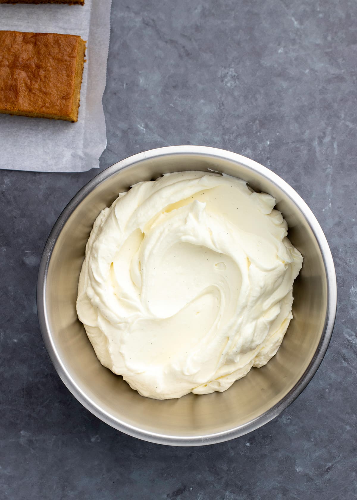 Cream cheese frosting in a large round metal bowl with plain pumpkin square in view
