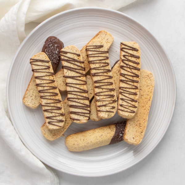 Closeup image of gluten free biscotti arranged in a white plate, with several pieces drizzled with melted chocolate and two pieces with one end dipped in chocolate and coarse sugar.