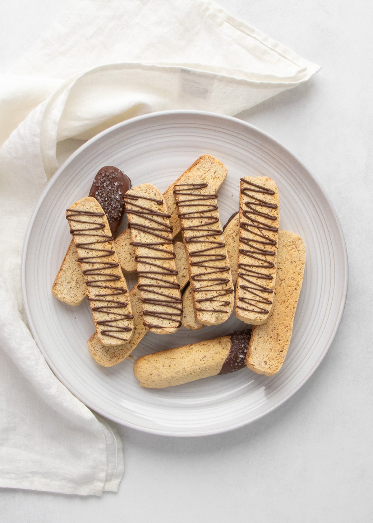 Overhead view of gluten free biscotti piled on a white plate, with several pieces drizzled with chocolate and two pieces partially dipped in chocolate, with a white cloth next to it.