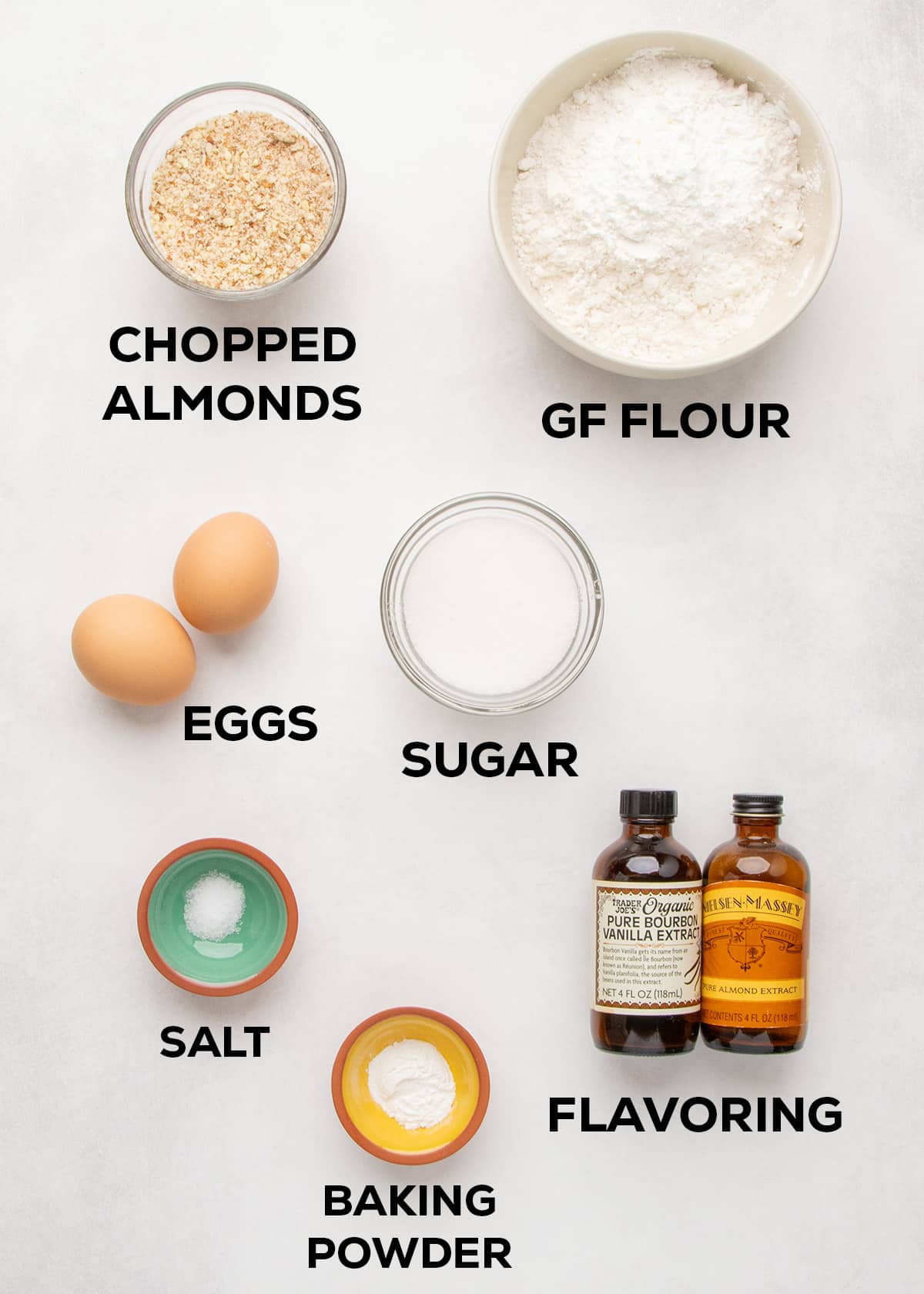 Chopped almonds, gluten free flour, eggs, sugar, salt, baking powder, vanilla extract, and almond extract arranged on a white surface.