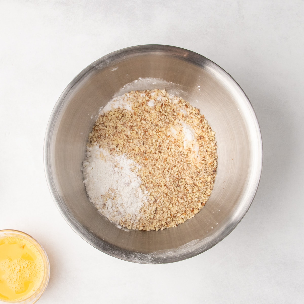 White flour mixture topped with finely chopped nuts in a stainless steel mixing bowl, with beaten egg partially visible at the lower left.
