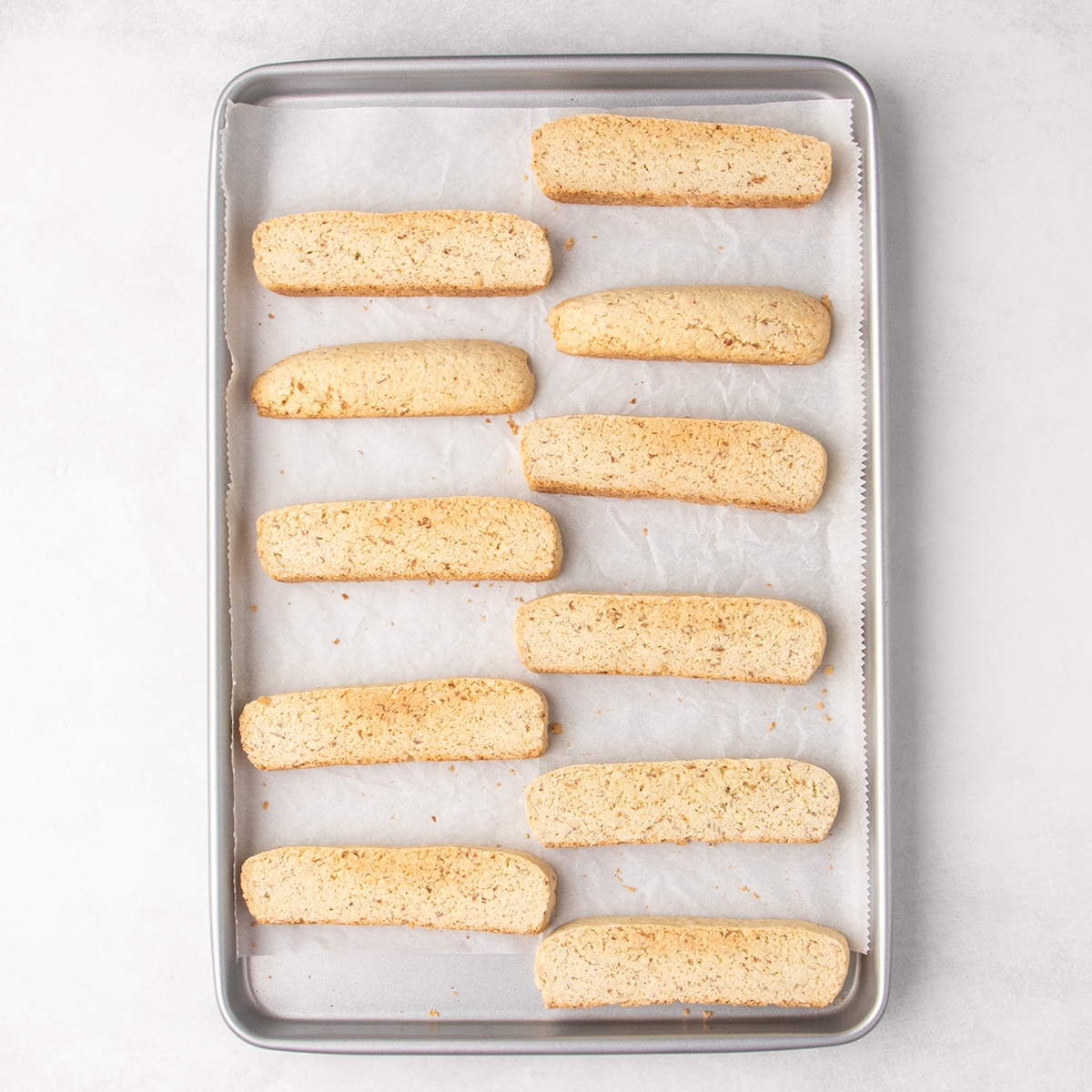 Eleven lightly browned fully baked gluten free biscotti pieces arranged cut-side up on a parchment-lined metal baking sheet.