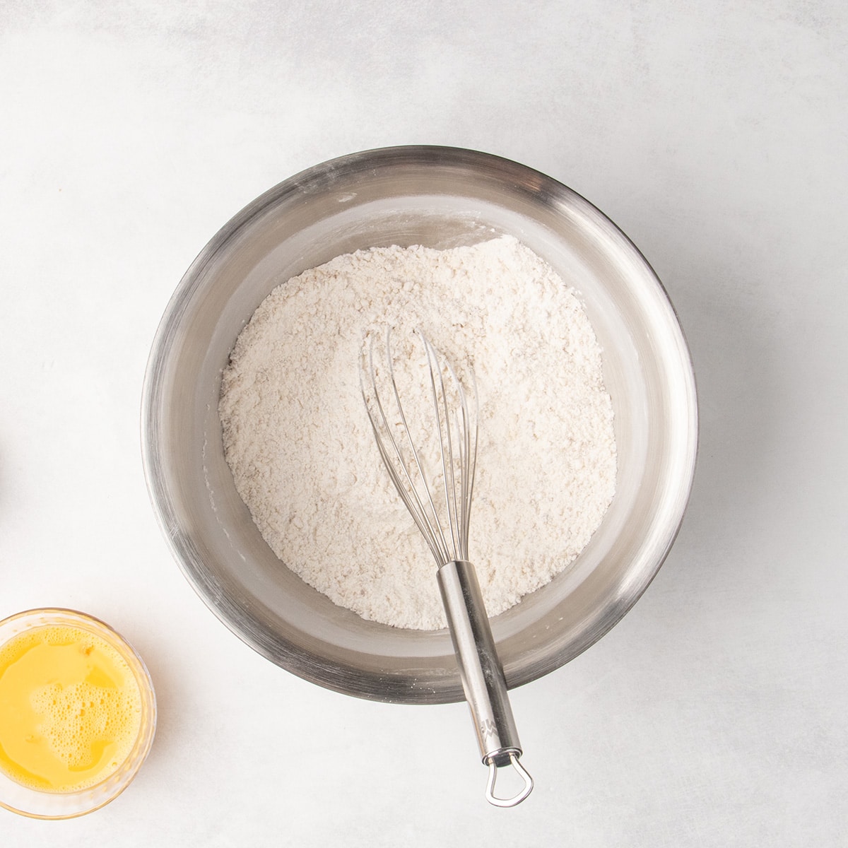 Pale dry ingredients whisked together in a stainless steel mixing bowl, with a metal whisk resting in the mixture.