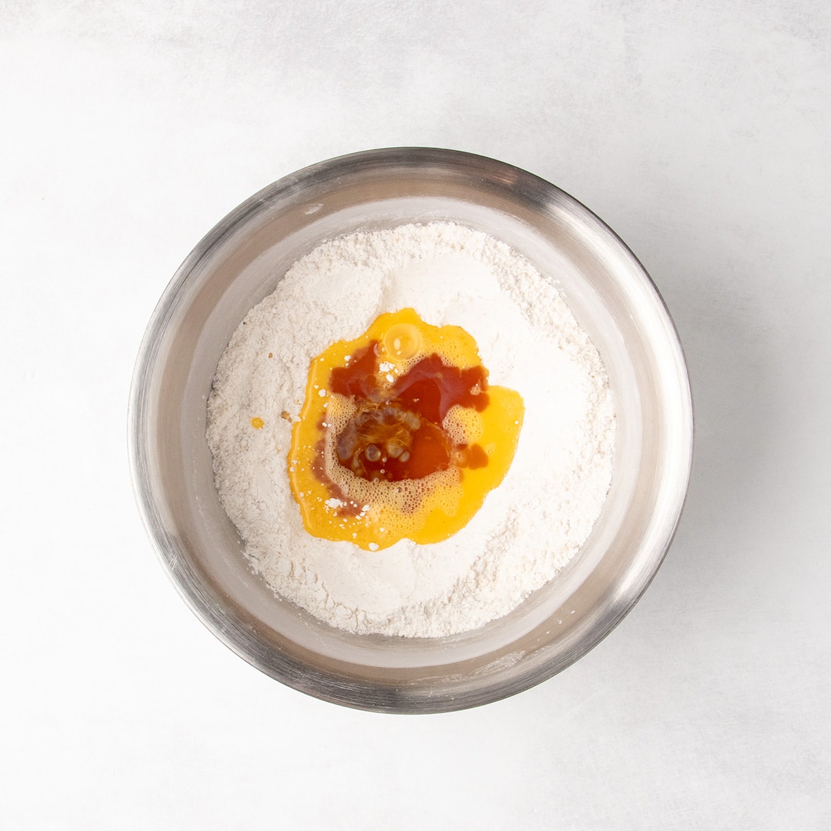 Yellow beaten egg and a dark amber liquid poured into a well in the dry ingredients in a stainless steel mixing bowl.