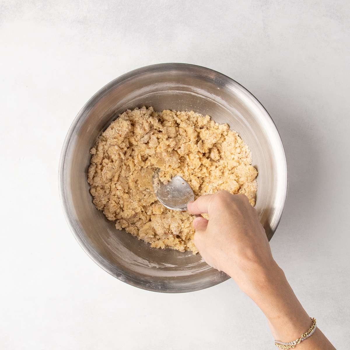 Hand pressing a metal mixing spoon into a crumbly pale dough in a stainless steel mixing bowl.