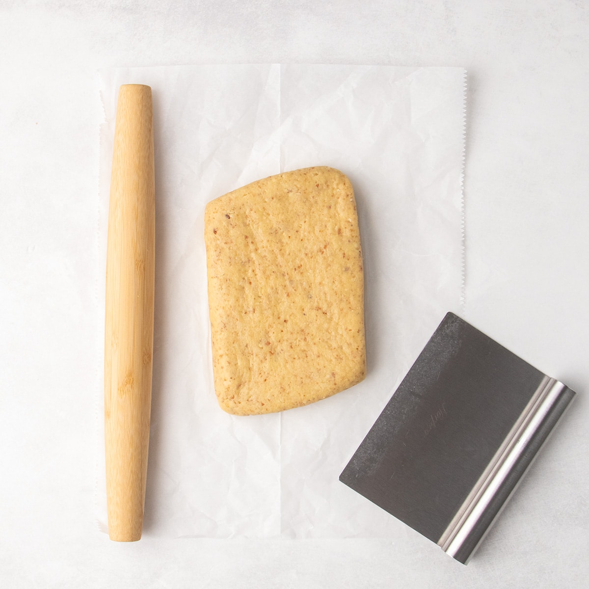 Flat slab of pale brown raw dough speckled with brown almond pieces on parchment paper shaped into a rectangle with angled short sides, and a wooden rolling pin on the left and a metal bench scraper on the right.