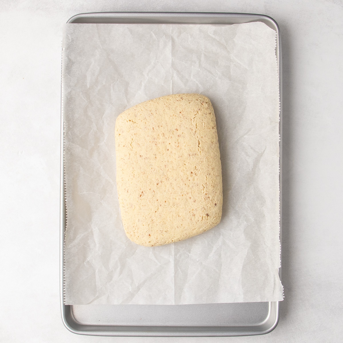 Once-baked flat loaf of very pale tan speckled dough centered on a parchment-lined metal baking sheet.