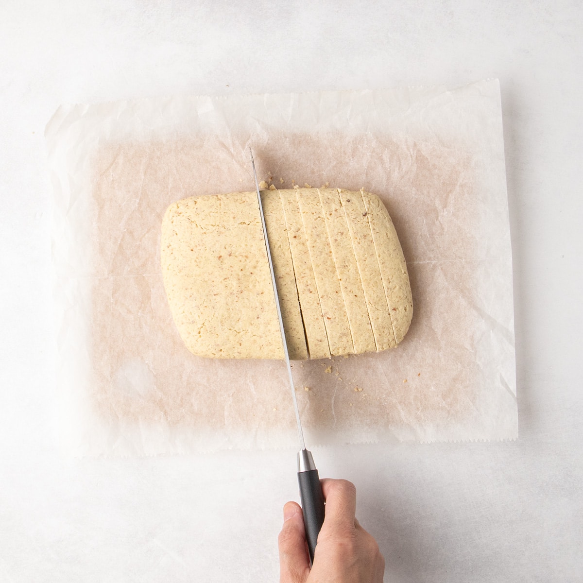 Hand holding knife slicing pale baked dough slab and cutting it crosswise into narrow slices.