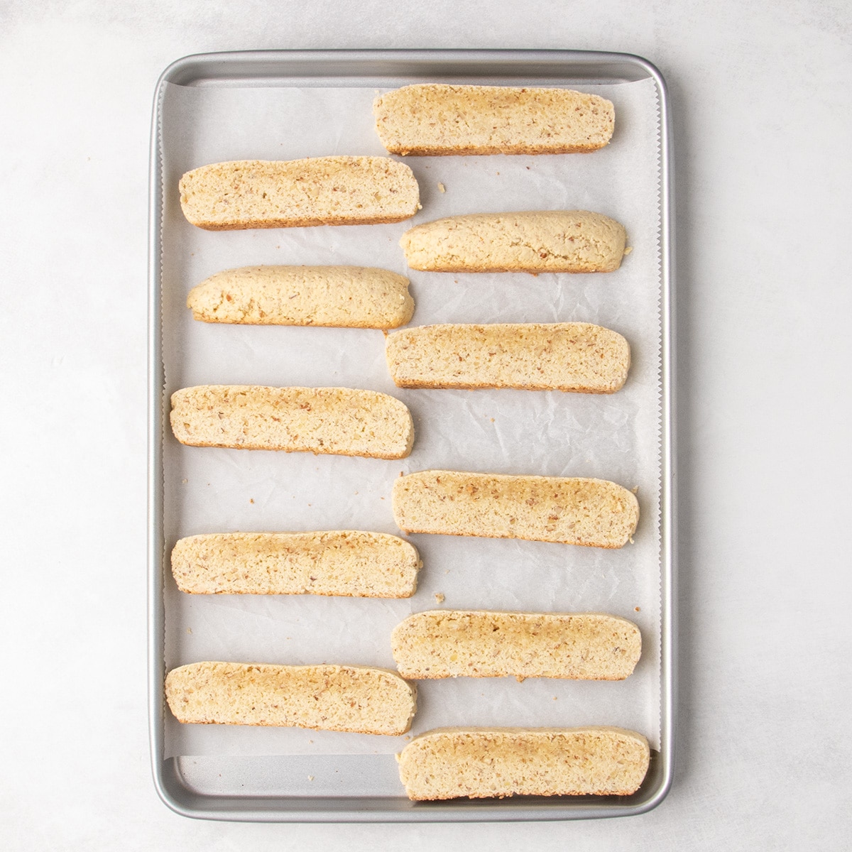 Eleven pale sliced partially baked biscotti pieces arranged cut-side up on a parchment-lined metal baking sheet.