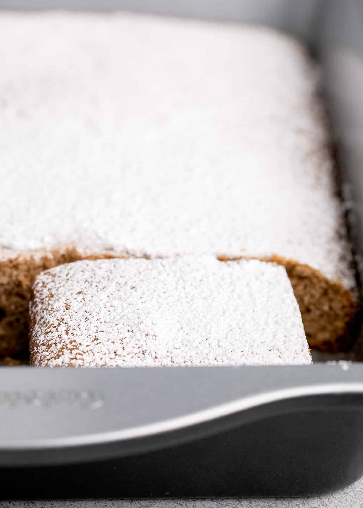 Powdered sugar on light brown spice cake in pan with a couple pieces missing