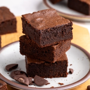 Closeup image of 3 thick chocolate protein brownies stacked on a brown-speckled white plate, with chopped chocolate pieces scattered around the base.