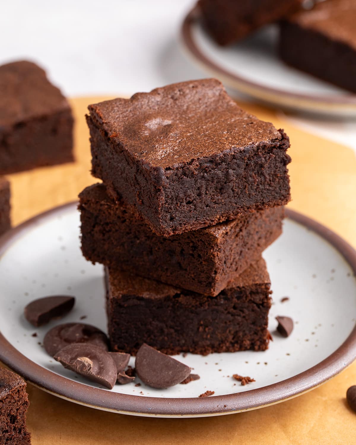 Three thick chocolate brownies stacked on a speckled plate, showing their dense centers and smooth tops, with more brownies on the side and background, mostly on a piece of brown paper