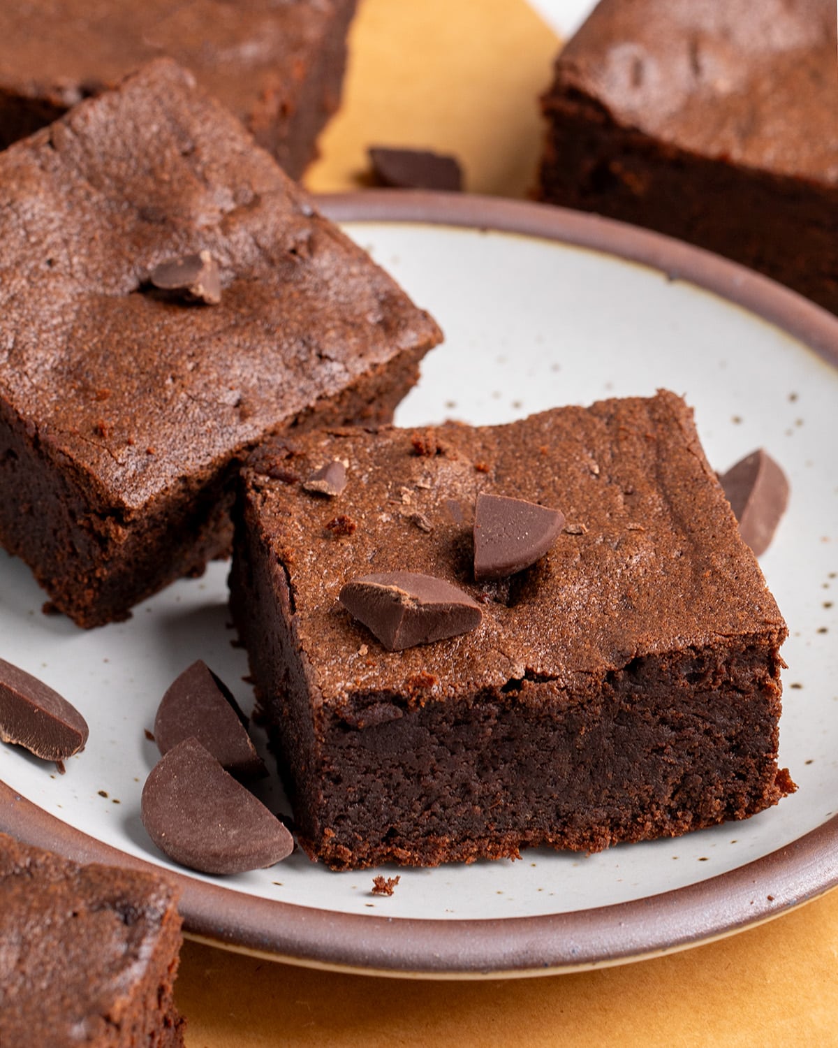 Close-up of two thick chocolate brownies on a speckled plate, topped and surrounded with broken chocolate pieces.