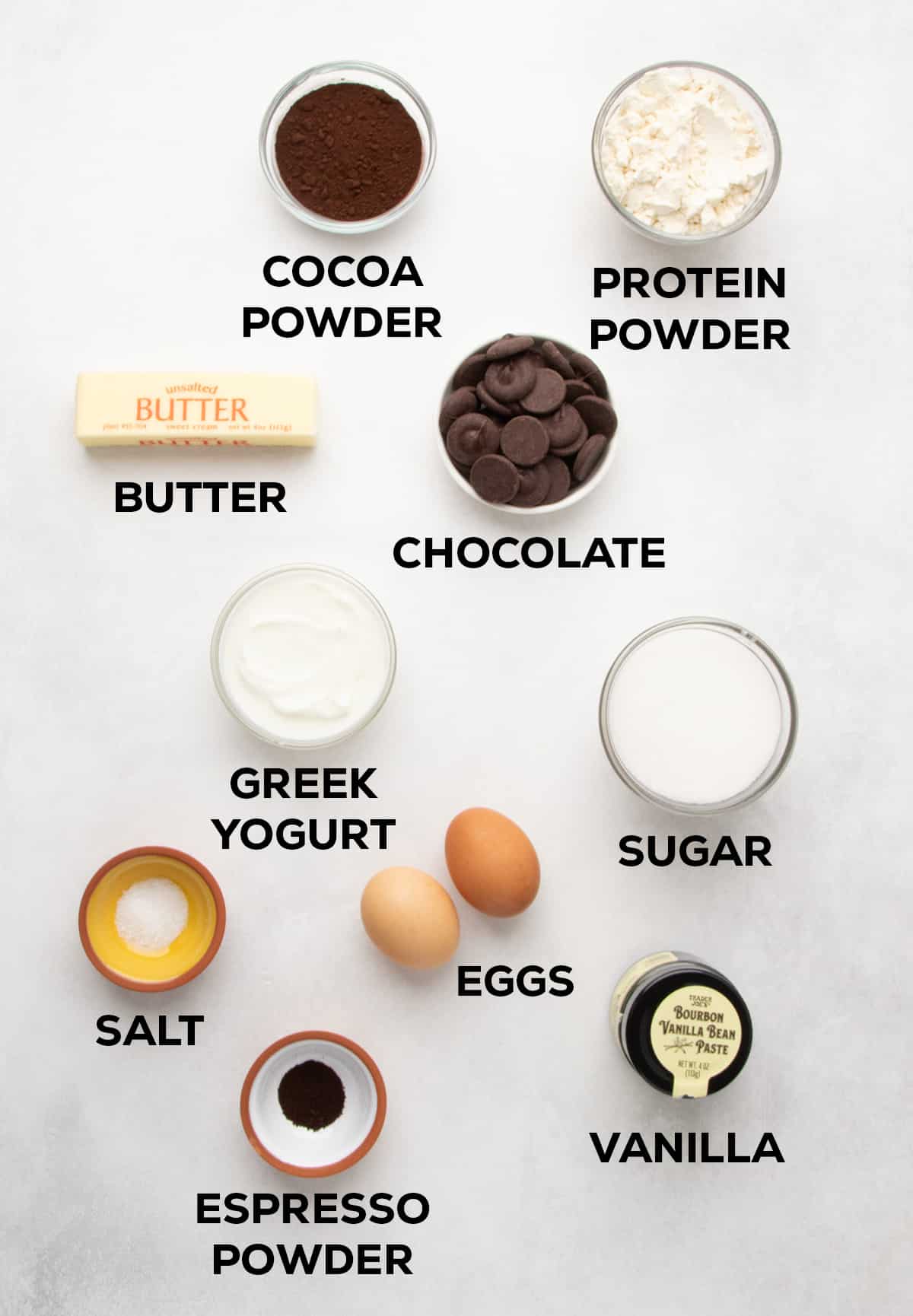Overhead arrangement of labeled ingredients including cocoa powder, protein powder, butter, chocolate, Greek yogurt, sugar, eggs, salt, espresso powder, and vanilla.