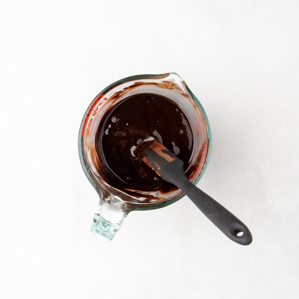 Overhead view of a glass measuring cup filled with dark, glossy melted chocolate and cocoa powder mixture and a black silicone spatula.