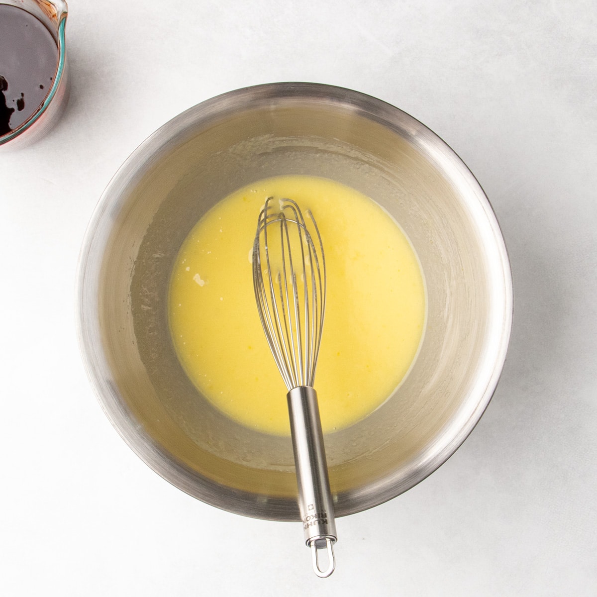Overhead view of a pale yellow liquid mixture in a stainless steel mixing bowl with a wire whisk resting inside.