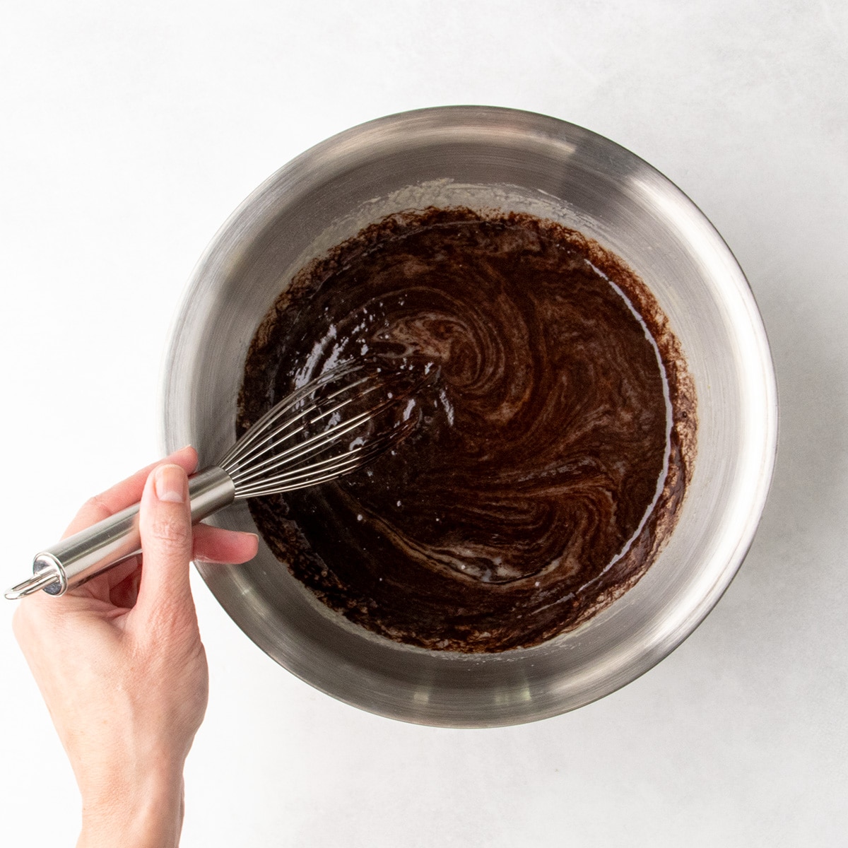 Hand whisking a dark brown chocolate mixture with pale yellow egg mixture in a stainless steel mixing bowl.