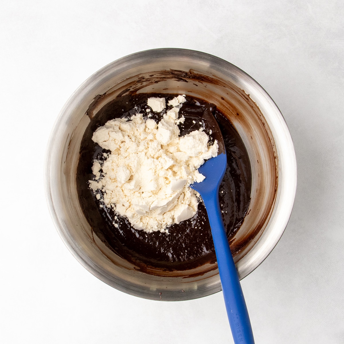 Pale protein powder piled over dark chocolate batter in a stainless steel bowl with a blue silicone spatula.