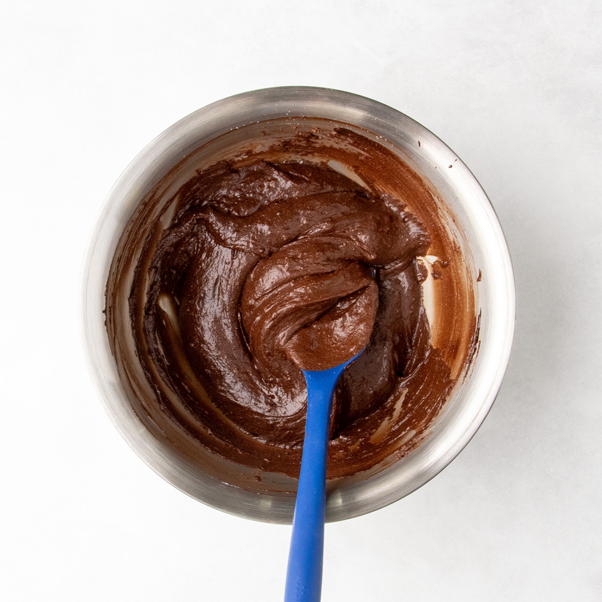 Thick, smooth chocolate brownie batter in a stainless steel mixing bowl with a blue silicone spatula.