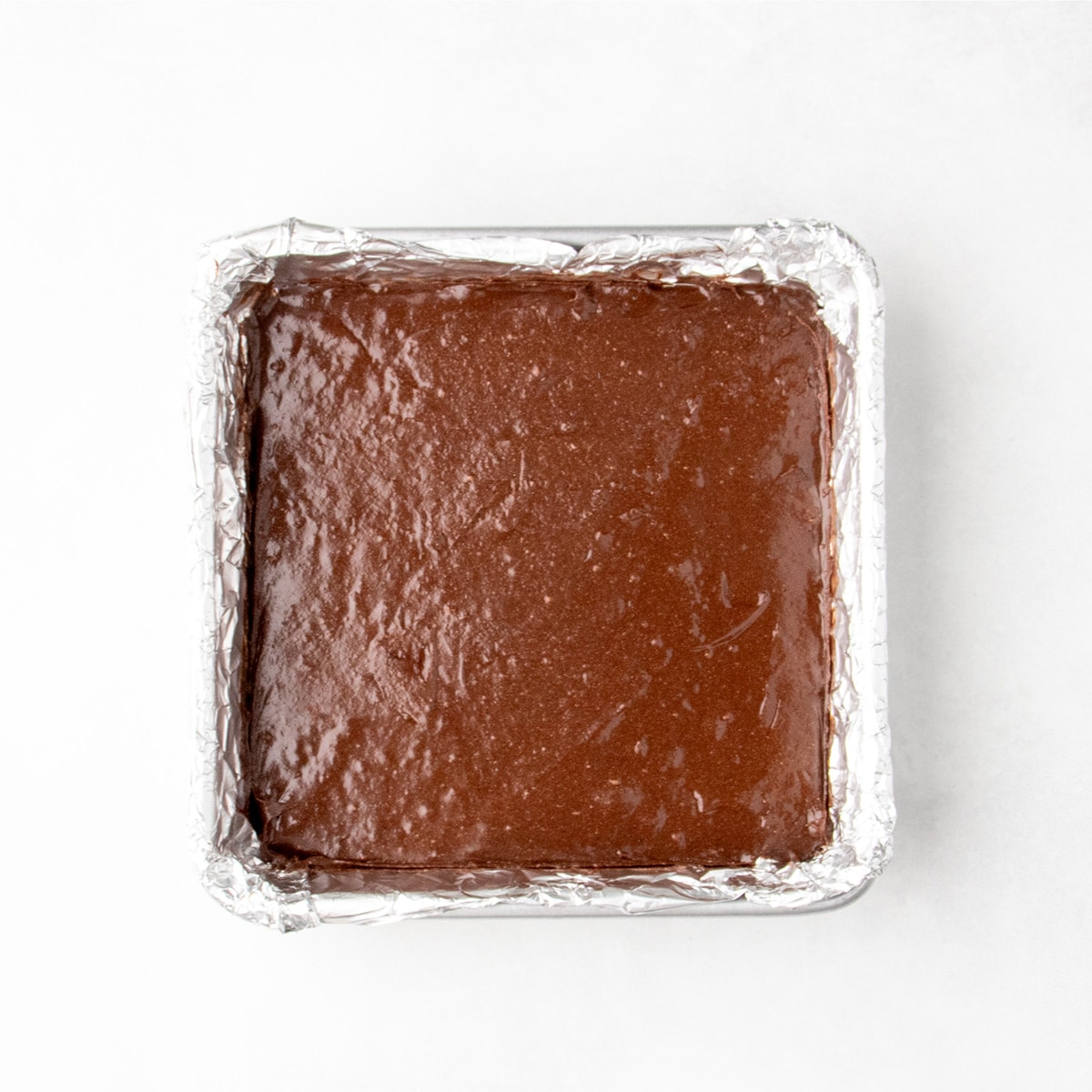 Smooth raw chocolate brownie batter spread evenly in a square foil-lined baking pan.