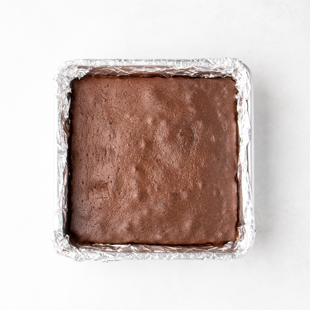 Baked chocolate brownies with a smooth dark brown top in a square foil-lined baking pan.