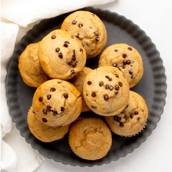Closeup image of protein muffins, some topped with mini chocolate chips, piled on a dark round tray.