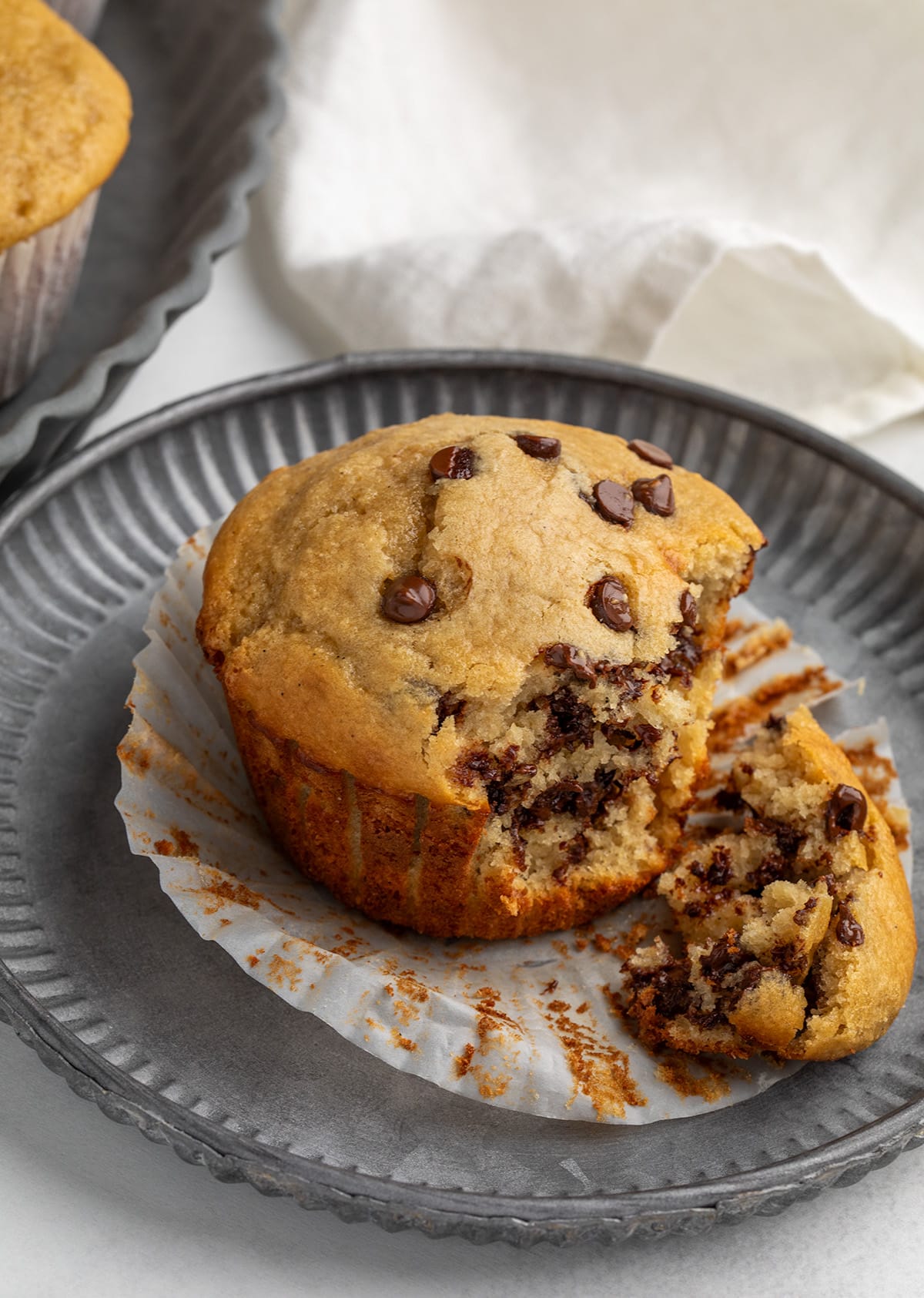A light brown muffin with mini chocolate chips is broken open on a dark plate, showing the soft interior and melted mini chocolate chips.
