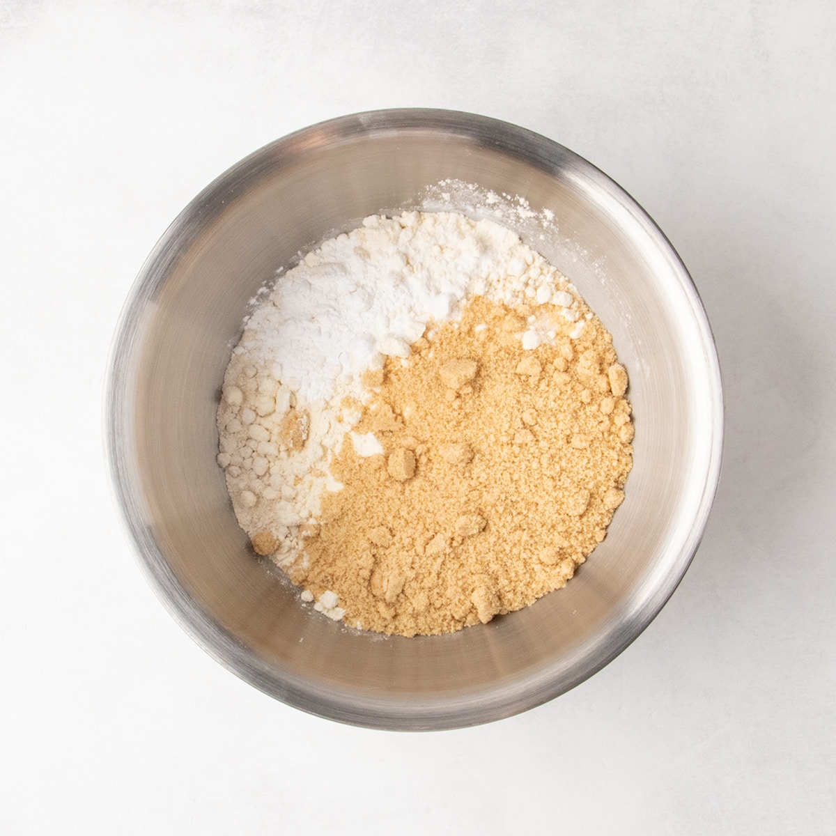 White and light brown dry ingredients are combined in a stainless steel mixing bowl.