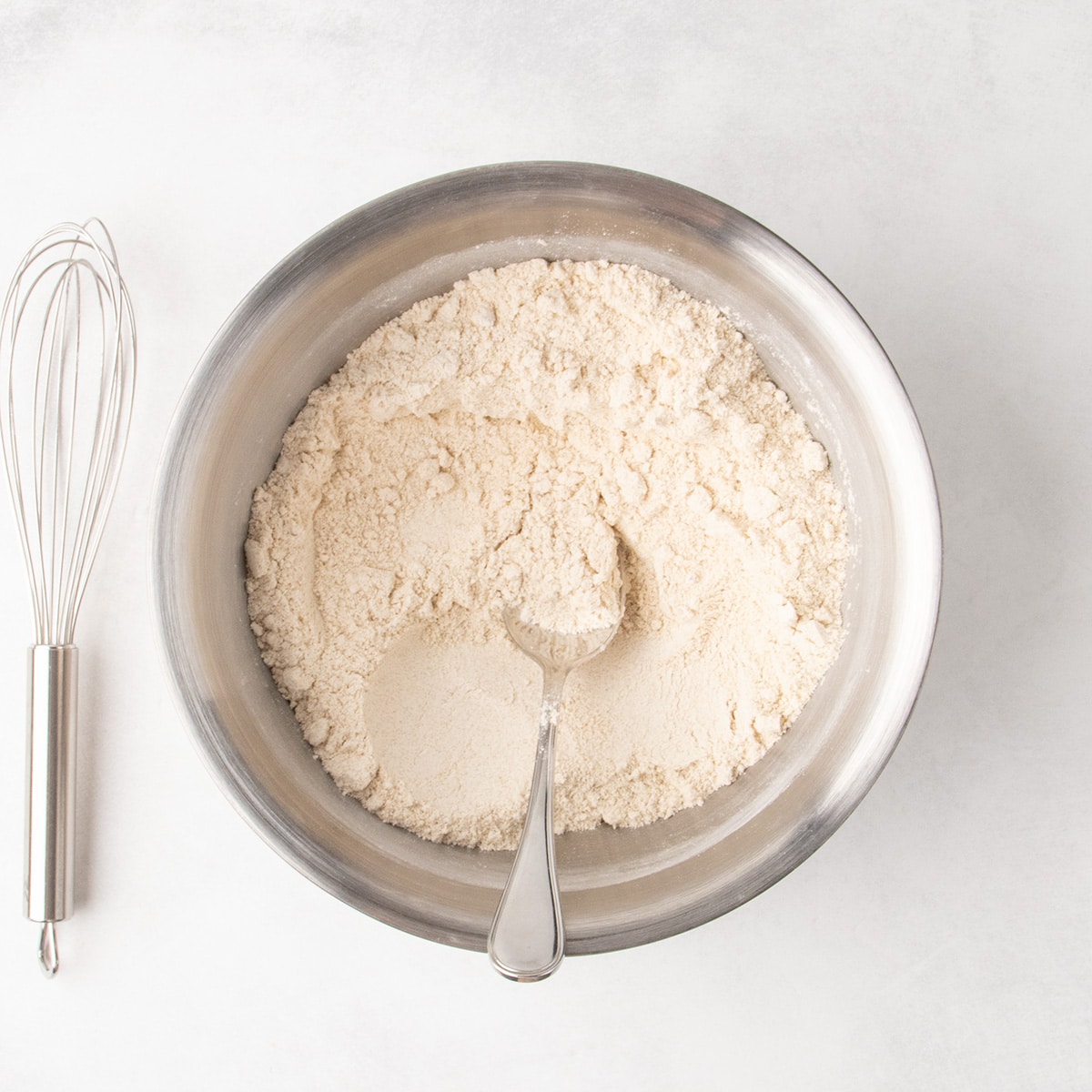 Very pale tan mixture of dry ingredients mixed together with a spoon in a stainless steel bowl, with a wire whisk next to the bowl.