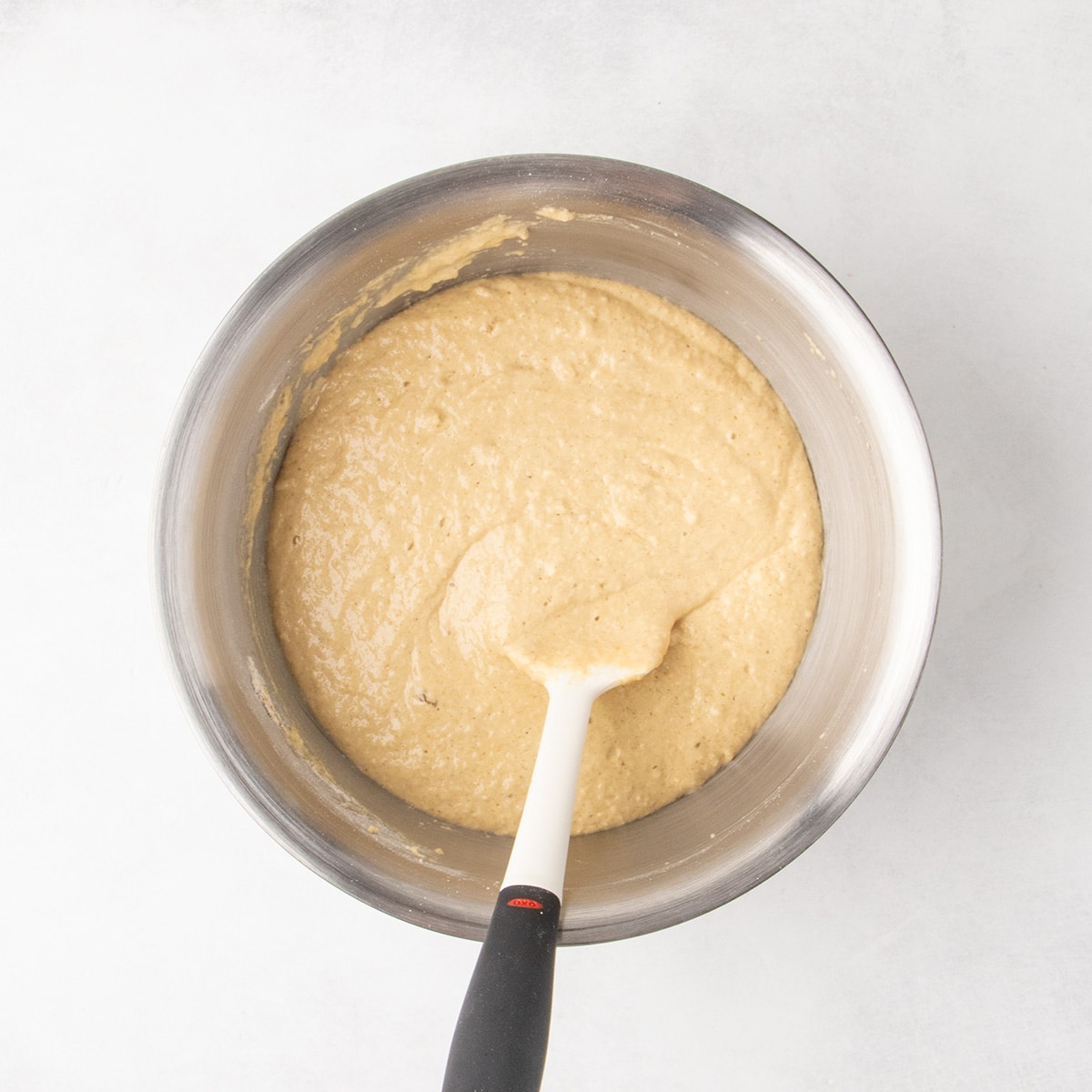 Pale brown, soft raw muffin batter mixed together with a white spatula with a black handle in a stainless steel bowl.