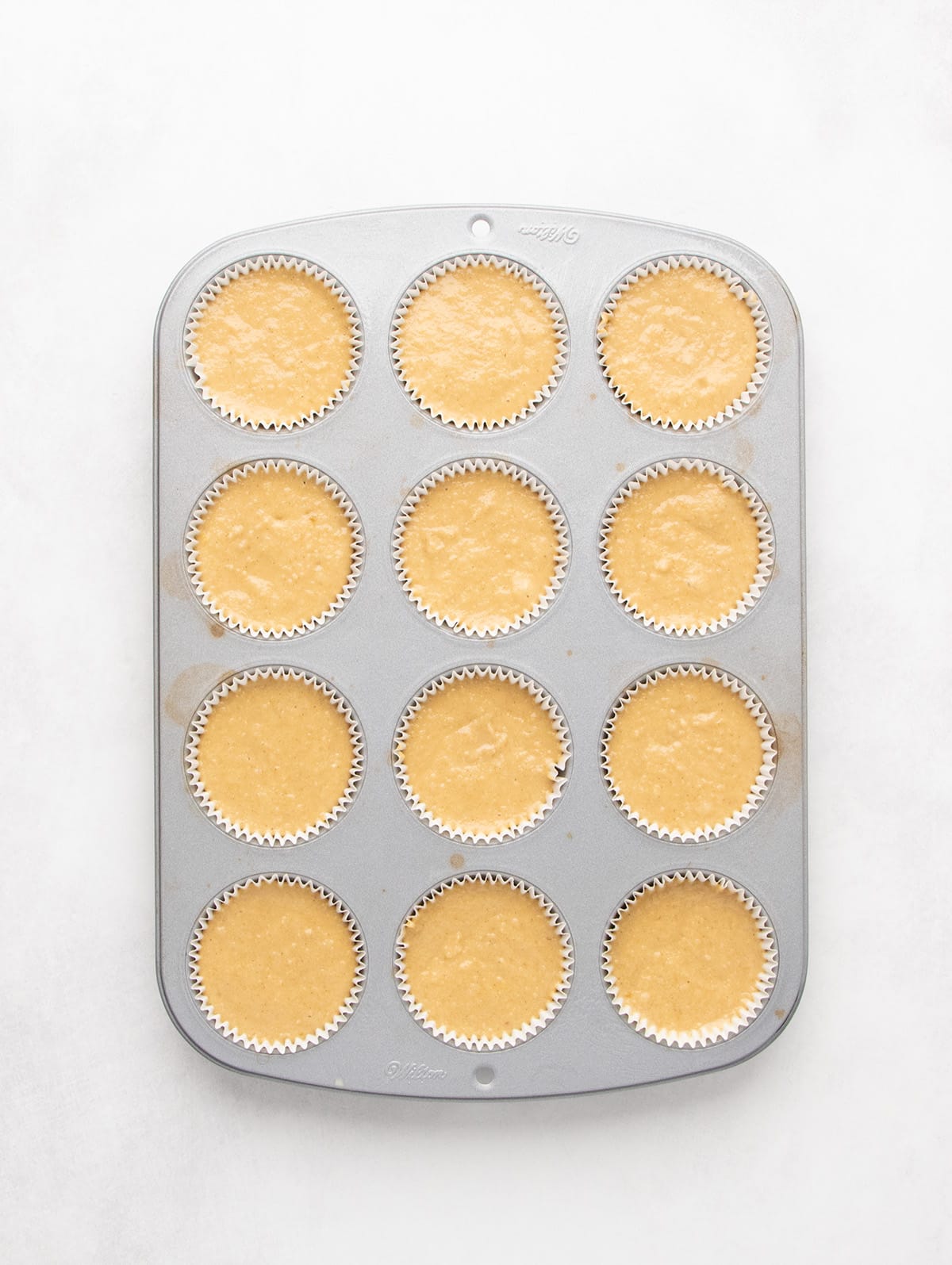 Light brown raw muffin batter is portioned into a lined cast aluminum 12-cup muffin tin.